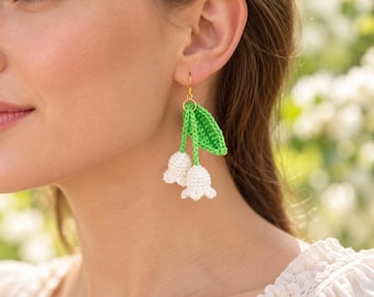 May include: A dangling earring featuring two small, white, crocheted bell-shaped flowers attached to a bright green crocheted leaf and stem. The piece is suspended from a gold-toned metal hook, showcasing intricate yarn texture and craftsmanship.
