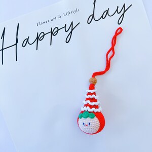May include: A red and white crocheted ornament with a smiling face and a green hat. The ornament is hanging from a red string and the text "Happy day" is written on a white background.