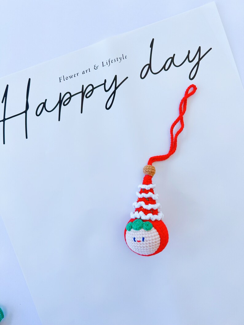 May include: A red and white crocheted ornament with a smiling face and a green hat. The ornament is hanging from a red string and the text "Happy day" is written on a white background.