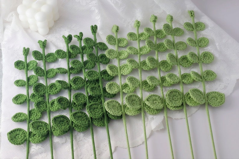 May include: Handmade crochet stems with green leaves. The stems are a light green color, and the leaves are a darker shade of green. The leaves are round and have a textured appearance. The stems are arranged in a row.