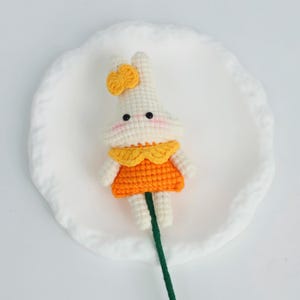 Puede incluir: Una mu&ntilde;eca de conejo de ganchillo hecha a mano con cuerpo blanco, vestido naranja y lazo amarillo. La mu&ntilde;eca tiene ojos negros y mejillas rosadas. Est&aacute; unida a un tallo verde, posiblemente para un ramo o decoraci&oacute;n.