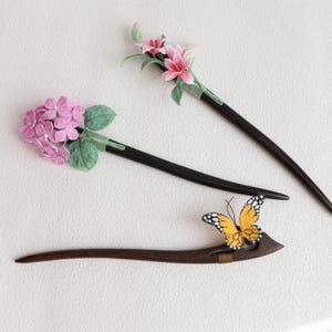 May include: Three decorative hair sticks. Two have dark brown stems with floral accents: pink lilies and pink hydrangea. The third stick is brown with a yellow and black butterfly accent. All are on a white background.