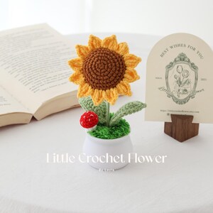 May include: A handcrafted crochet sunflower in a white pot, with green leaves and a red mushroom. The sunflower has a yellow and brown color scheme. The text "Little Crochet Flower" is visible.