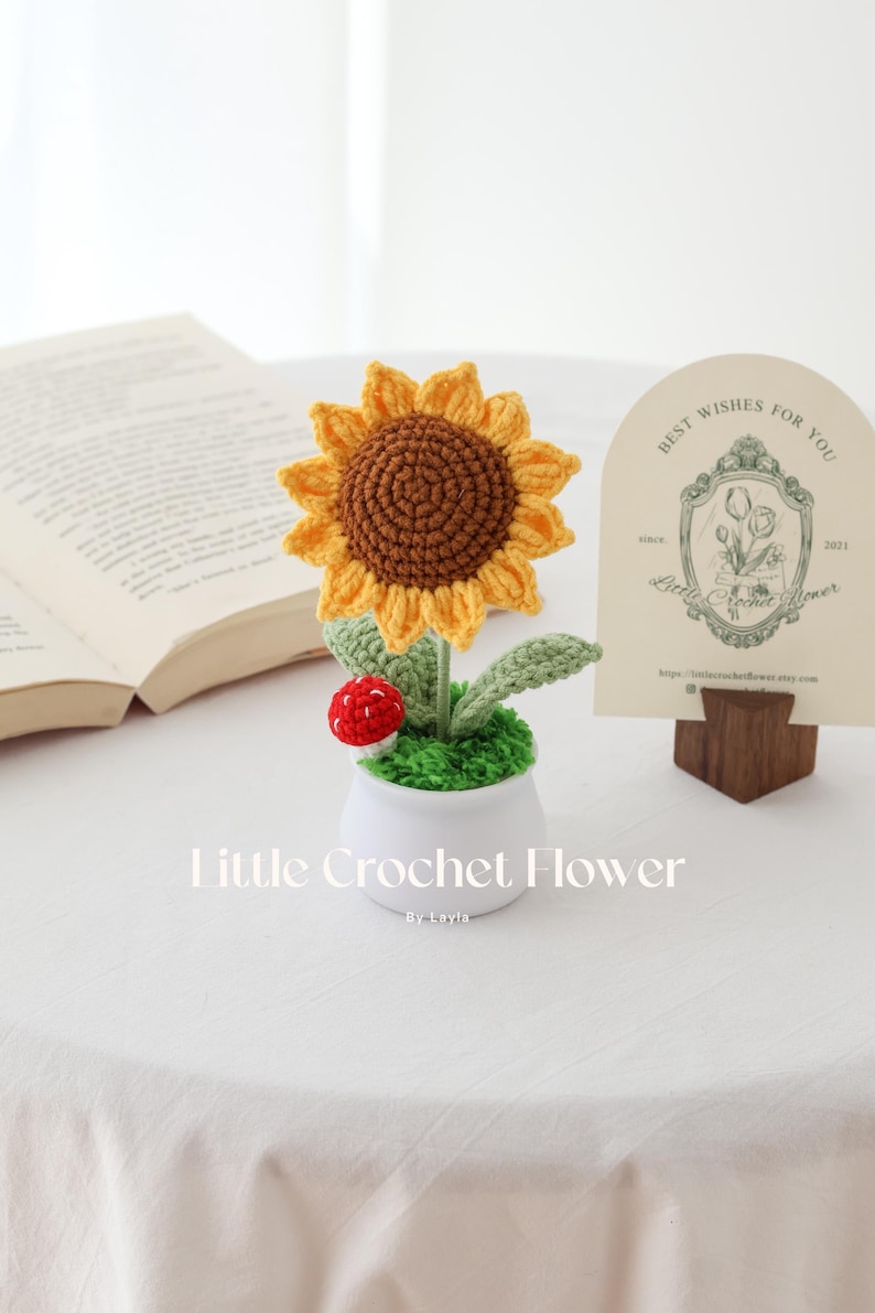May include: A handcrafted crochet sunflower in a white pot, with green leaves and a red mushroom. The sunflower has a yellow and brown color scheme. The text "Little Crochet Flower" is visible.