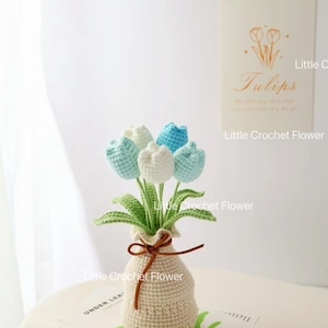 May include: A handmade crochet bouquet of tulips in a beige vase. The flowers are white and light blue, with green leaves. The vase is tied with a brown string. The text "Little Crochet Flower" is visible.