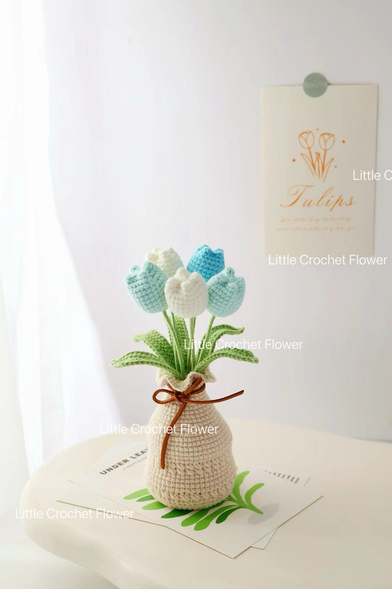 May include: A handmade crochet bouquet of tulips in a beige vase. The flowers are white and light blue, with green leaves. The vase is tied with a brown string. The text "Little Crochet Flower" is visible.