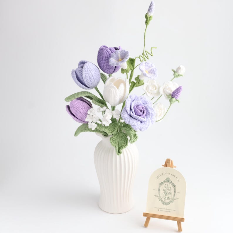 May include: A bouquet of crocheted flowers in shades of lavender, purple, and white, featuring tulips, roses, and small blossoms with green stems and leaves, arranged in a white fluted vase next to a small wooden easel holding a card with text.