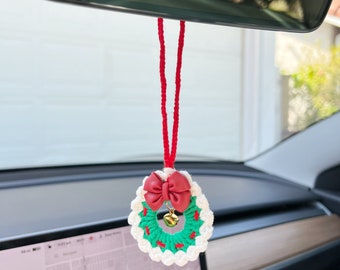 May include: A crocheted Christmas wreath car charm with a red bow and a small gold bell. The wreath is made with green and white yarn, suspended from a red string, and hangs from a car's rearview mirror.