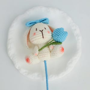 Puede incluir: Un conejito de ganchillo hecho a mano, blanco con detalles rosas, con un lazo azul y una flor de tulip&aacute;n azul. El tulip&aacute;n tiene un tallo y hojas verdes. El conejito est&aacute; presentado sobre un plato blanco, ideal para un regalo.