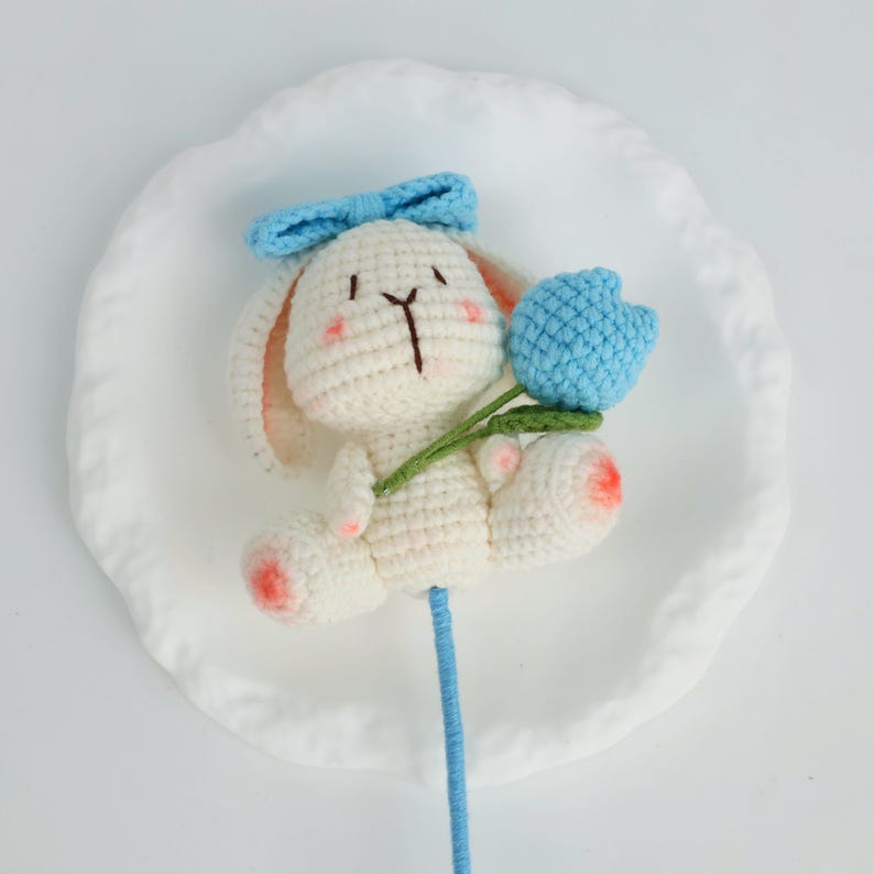 Puede incluir: Un conejito de ganchillo hecho a mano, blanco con detalles rosas, con un lazo azul y una flor de tulip&aacute;n azul. El tulip&aacute;n tiene un tallo y hojas verdes. El conejito est&aacute; presentado sobre un plato blanco, ideal para un regalo.