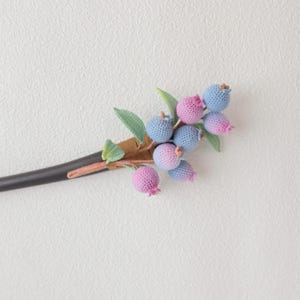 May include: A handcrafted hair stick featuring a cluster of crocheted blueberries in shades of blue and pink. The berries are attached to a brown stem with green leaves, and the stick itself is black.