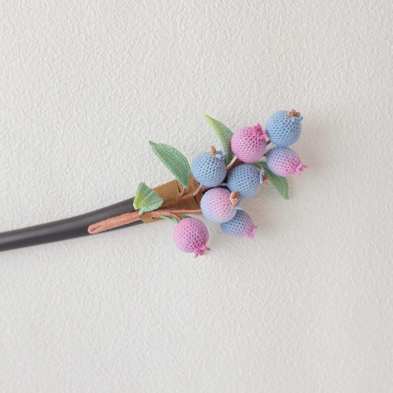 May include: A handcrafted hair stick featuring a cluster of crocheted blueberries in shades of blue and pink. The berries are attached to a brown stem with green leaves, and the stick itself is black.