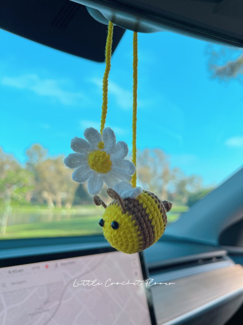 May include: A crocheted bee with brown and yellow stripes and black eyes hangs from a rearview mirror in a car. The bee is holding a white crocheted daisy with a yellow center.