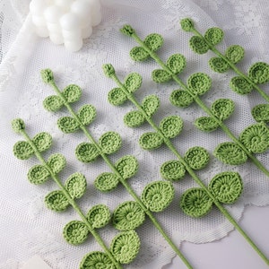 May include: Crocheted green leaf stems arranged on a white lace surface. The leaves are a light green color and have a detailed, textured appearance. A white bubble candle is in the background. The stems are likely for crafting or home decor.