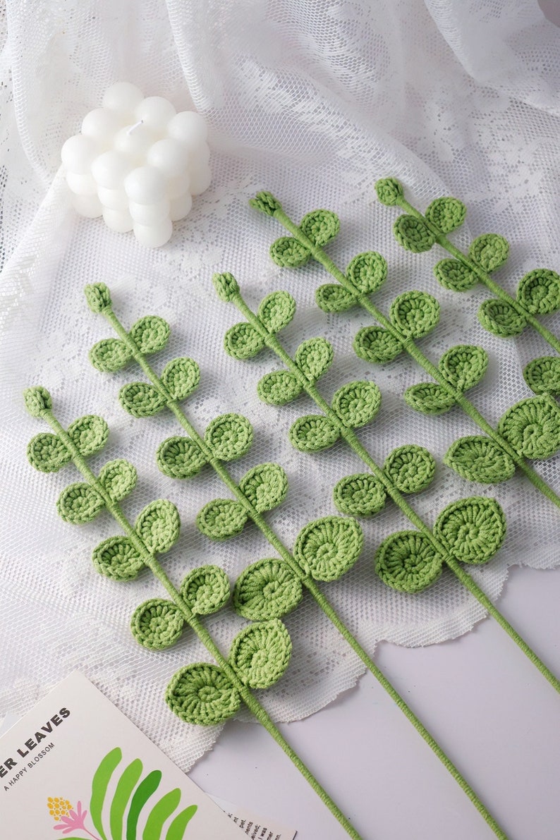 May include: Crocheted green leaf stems arranged on a white lace surface. The leaves are a light green color and have a detailed, textured appearance. A white bubble candle is in the background. The stems are likely for crafting or home decor.