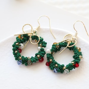 May include: A pair of green Christmas wreath earrings with gold accents and colorful beads. The earrings are made of crocheted yarn and feature a small bow at the top.
