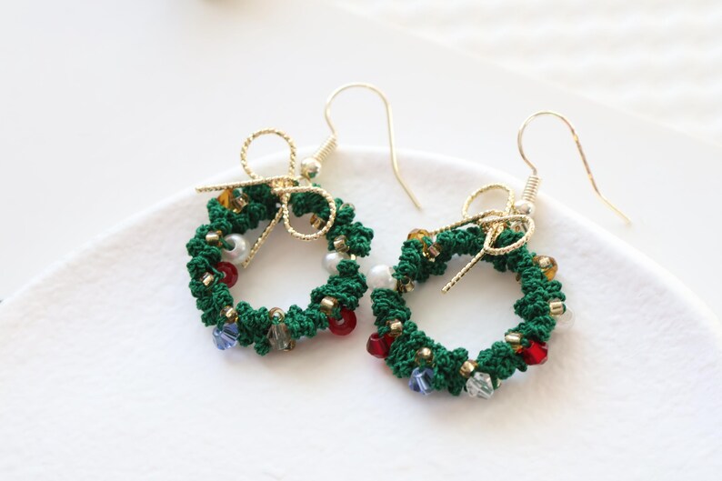 May include: A pair of green Christmas wreath earrings with gold accents and colorful beads. The earrings are made of crocheted yarn and feature a small bow at the top.