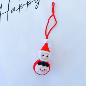 May include: A red and white crocheted Santa Claus ornament with a black string hanging from the top. The ornament has a white beard and a black moustache.