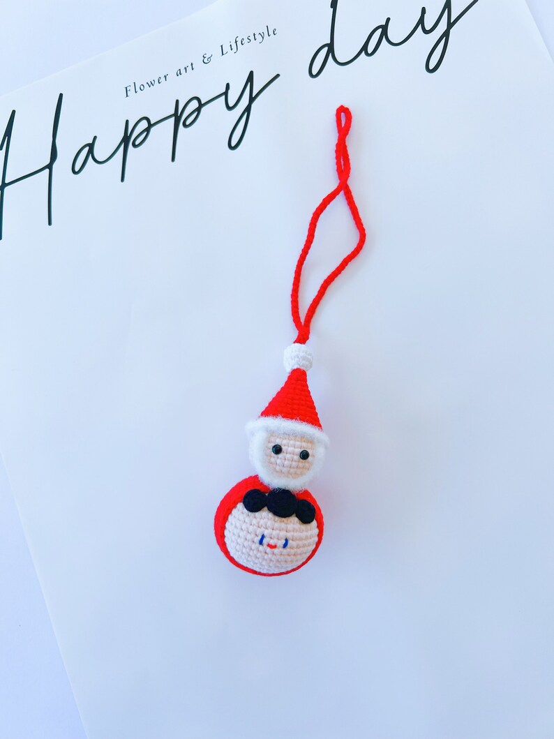May include: A red and white crocheted Santa Claus ornament with a black string hanging from the top. The ornament has a white beard and a black moustache.