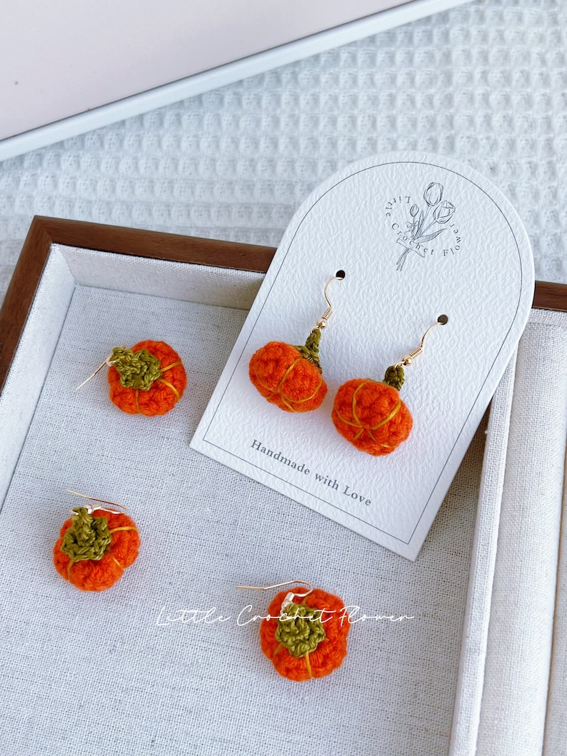 May include: Crocheted pumpkin earrings with green stems and gold hooks. The earrings are displayed on a white card with the text "Handmade with Love" and "Little Crochet Flower".