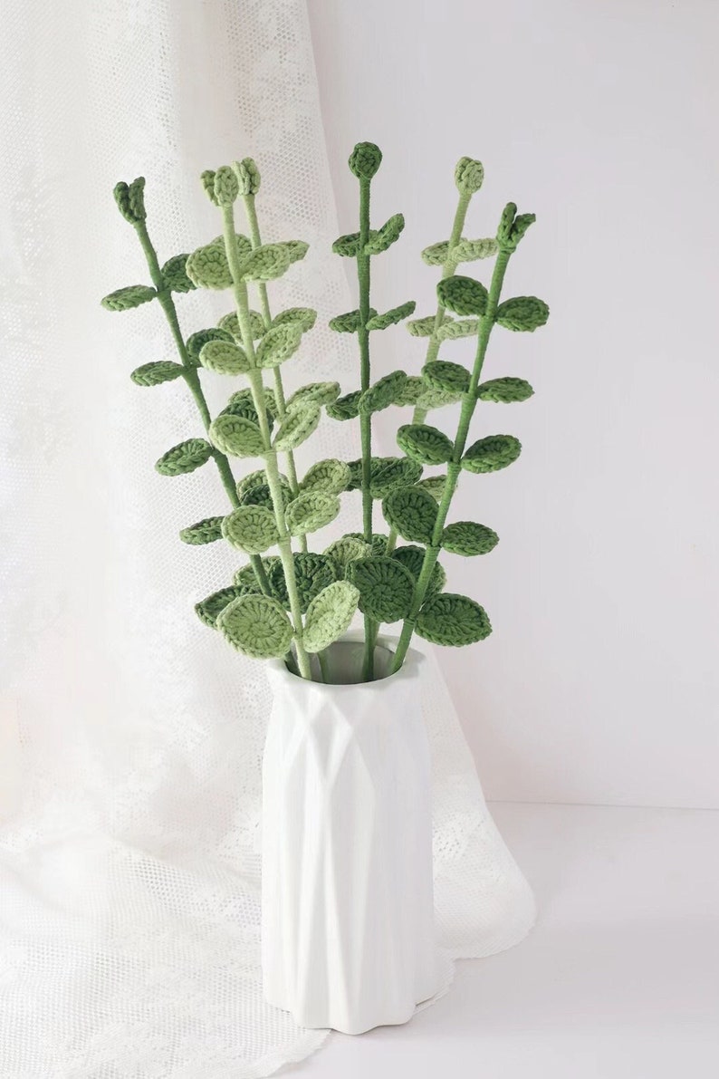 May include: A white ceramic vase holds a collection of handmade crochet stems with green leaves. The stems are a light green color, and the leaves are a darker shade of green. The vase has a geometric design.