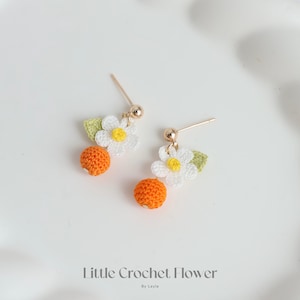 May include: A pair of crocheted earrings featuring a small orange sphere and a white flower with a yellow center. The earrings are attached to gold posts.