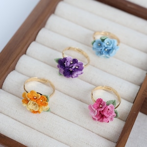 Può includere: Cinque anelli fatti a mano con disegni floreali in un espositore in legno. Gli anelli presentano fasce dorate e fiori all'uncinetto in vari colori: arancione, viola, rosa, blu e giallo. Ogni fiore ha un design unico e piccoli dettagli di perline.