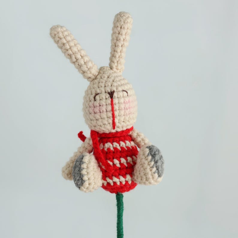 Puede incluir: Un juguete de conejo tejido a crochet con orejas largas, un chaleco con estampado rojo y blanco y patas grises. El conejo tiene una cara sonriente con mejillas rosadas y una nariz roja. El juguete est&aacute; unido a un tallo verde.