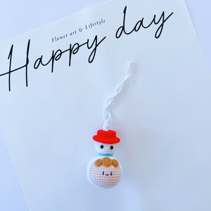 May include: A white crocheted snowman ornament with a red hat and a brown yarn face. The ornament is hanging from a white string and has the text "Happy day" written in black cursive on a white background.