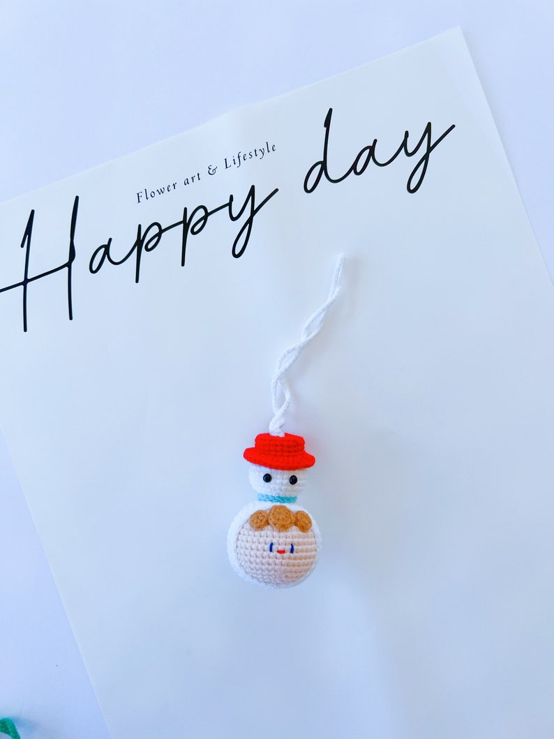 May include: A white crocheted snowman ornament with a red hat and a brown yarn face. The ornament is hanging from a white string and has the text "Happy day" written in black cursive on a white background.