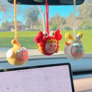 Puede incluir: Tres adornos colgantes de crochet en un coche. Los adornos son un cangrejo amarillo, un cordero amarillo y un cangrejo rojo. Los adornos cuelgan del espejo retrovisor.