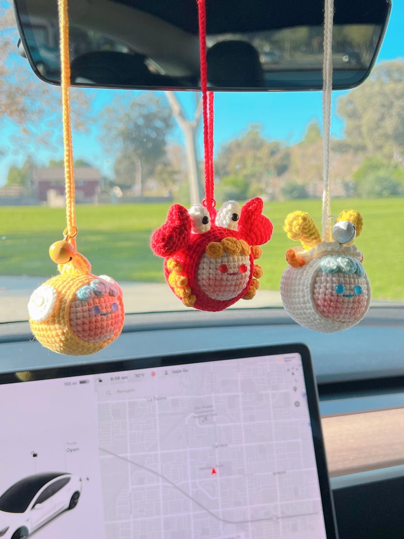 Puede incluir: Tres adornos colgantes de crochet en un coche. Los adornos son un cangrejo amarillo, un cordero amarillo y un cangrejo rojo. Los adornos cuelgan del espejo retrovisor.