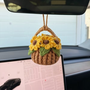May include: A crocheted car charm shaped like a basket of sunflowers. The basket is brown with a green rim and handle, and it is filled with yellow sunflowers with brown centers and green leaves. The charm hangs from a brown string.