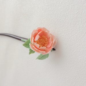 May include: A handcrafted hair accessory featuring a peach-colored flower with a detailed, textured design. The flower has a yellow center and is accompanied by green leaves. The accessory is attached to a dark brown stick, likely for insertion into the hair.