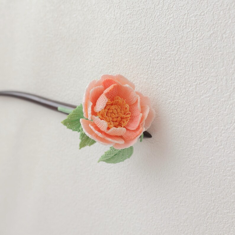 May include: A handcrafted hair accessory featuring a peach-colored flower with a detailed, textured design. The flower has a yellow center and is accompanied by green leaves. The accessory is attached to a dark brown stick, likely for insertion into the hair.