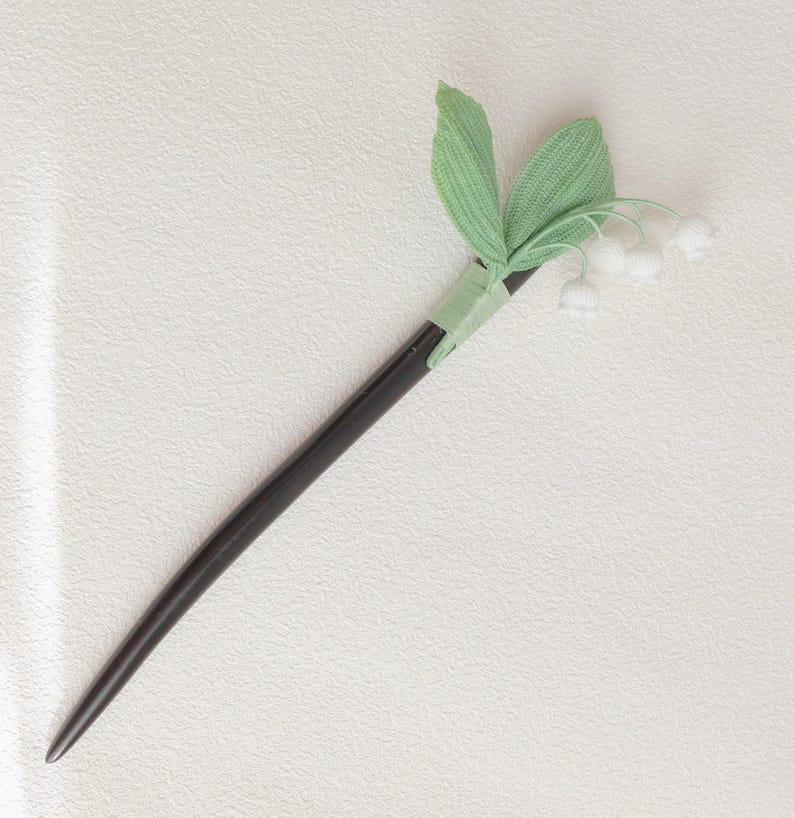 May include: A dark brown wooden hair stick with a decorative lily of the valley design. The hair accessory features light green leaves and small white flowers, adding a touch of nature-inspired elegance.