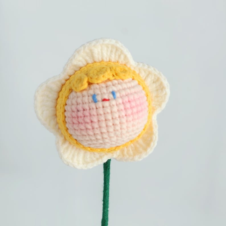 Puede incluir: Una flor de ganchillo hecha a mano con una cara sonriente. La flor tiene un dise&ntilde;o de p&eacute;talos blancos, un centro amarillo y un tallo verde. La cara tiene ojos azules, boca roja y mejillas rosadas. Un art&iacute;culo decorativo caprichoso.