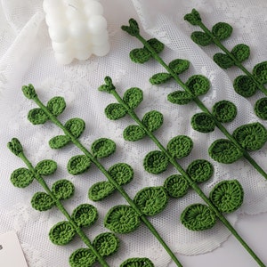 May include: Crocheted green leaf stems arranged on a white lace surface. The stems have small, round leaves and are a vibrant green color. A white bubble candle is in the background.