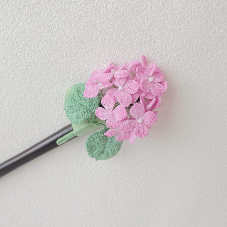May include: A handcrafted hair accessory featuring a cluster of pink hydrangea flowers with white centers and green leaves. The flowers are attached to a black stick, creating a delicate and floral design.