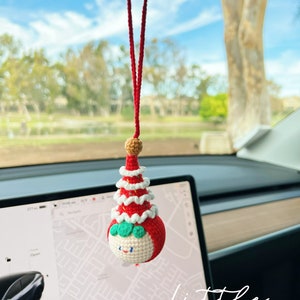 May include: A red and white crocheted Christmas tree ornament with a smiling face and green eyes. The ornament is hanging from a red string in a car.