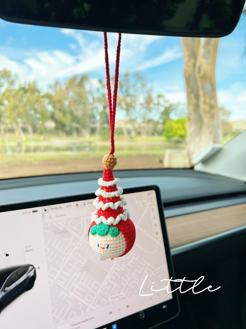 May include: A red and white crocheted Christmas tree ornament with a smiling face and green eyes. The ornament is hanging from a red string in a car.
