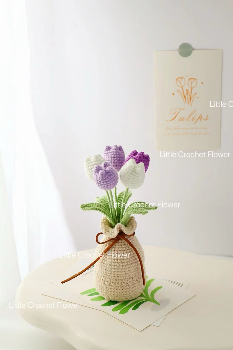 May include: A crocheted bouquet of tulips in a beige vase. The flowers are white, lavender, and purple, with green leaves. The vase is tied with a brown leather cord. A card with the word "Tulips" is in the background.