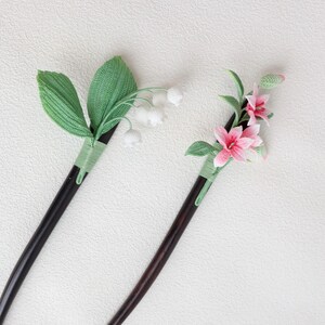 May include: Two decorative hair sticks. One features white lily of the valley flowers and green leaves. The other has pink lilies and green leaves. Both have dark brown wooden stems and are wrapped with green thread.