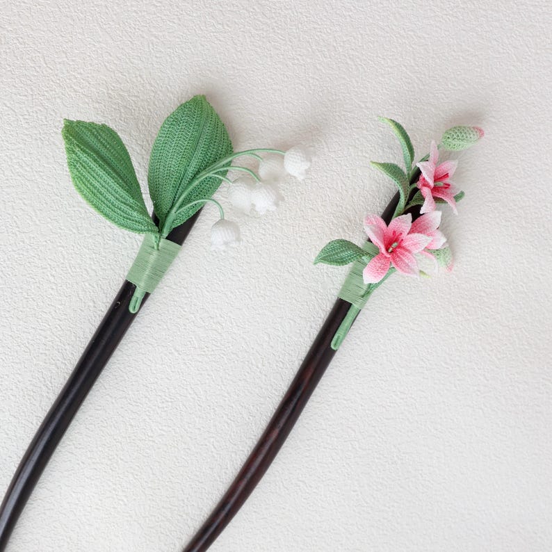 May include: Two decorative hair sticks. One features white lily of the valley flowers and green leaves. The other has pink lilies and green leaves. Both have dark brown wooden stems and are wrapped with green thread.