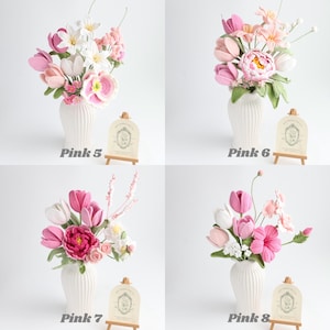 May include: Four arrangements of crocheted flowers in white ribbed vases, labeled Pink 5, Pink 6, Pink 7, and Pink 8. Each bouquet features various shades of pink, white, and green yarn-crafted blossoms, buds, and leaves with small wooden display signs.