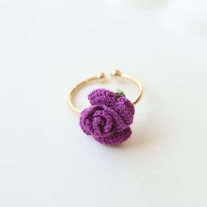 May include: A purple crocheted flower ring with a gold band. The flower is attached to the band with a small green stem.