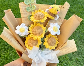 May include: A bouquet of crocheted flowers, including sunflowers, daisies, and a bee, wrapped in brown paper and tied with a white ribbon. The bouquet has a green stem and leaves. The text "Little Crochet Flower" is visible.