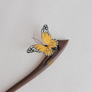 May include: A wooden hair stick with a decorative yellow and black butterfly. The butterfly has detailed wing patterns and is attached to the dark brown, curved stick. The stick has a small gold band near the top.