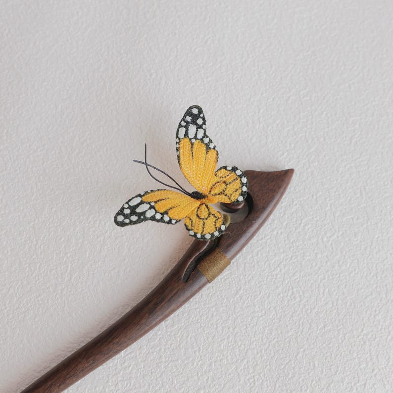 May include: A wooden hair stick with a decorative yellow and black butterfly. The butterfly has detailed wing patterns and is attached to the dark brown, curved stick. The stick has a small gold band near the top.