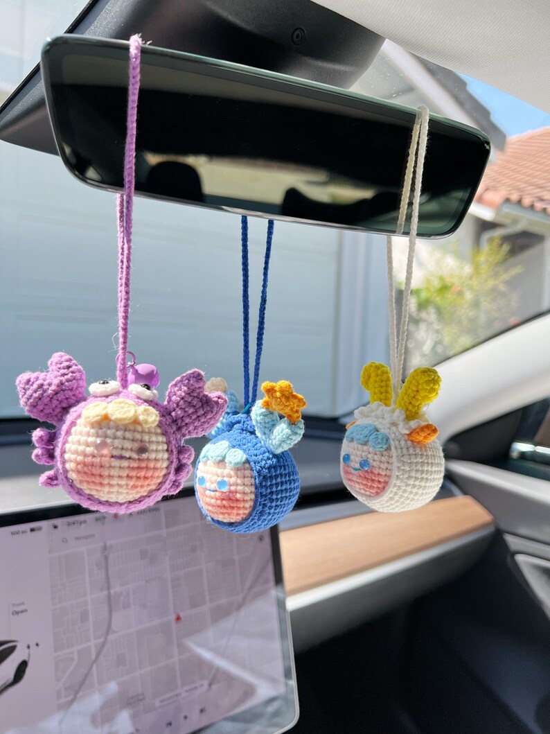 Puede incluir: Tres adornos de animales del zod&iacute;aco hechos a crochet colgando del espejo retrovisor de un coche. Los adornos son un cangrejo morado, un pez azul y un carnero blanco.